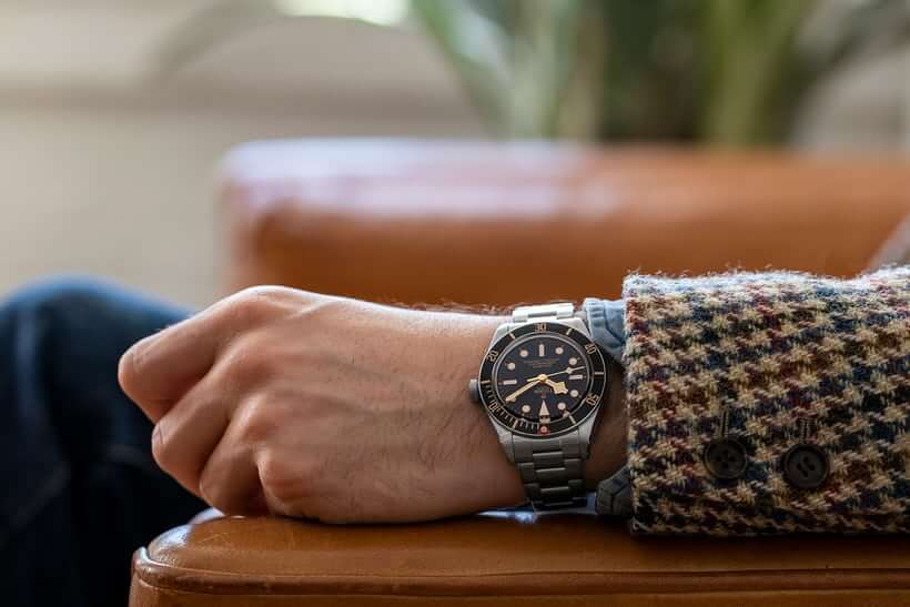 Seorang pria dengan setelan jas mengenakan jam tangan stainless steel sedang duduk di kursi kayu